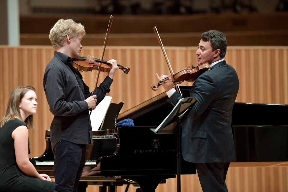 A male violin professor, holding a violin, teaching a male student, holding a violin, with a female student sitting by a piano.